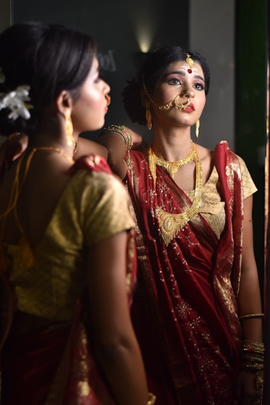 A woman adorned in traditional Indian attire is gazing at her reflection in the mirror. She wears a vibrant red saree with gold embroidery and heavy traditional jewelry, including a nose ring and a headpiece. Her makeup is meticulously done, emphasizing her eyes and lips, and she has flowers tucked into her neatly styled hair.