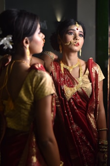 A woman adorned in traditional Indian attire is gazing at her reflection in the mirror. She wears a vibrant red saree with gold embroidery and heavy traditional jewelry, including a nose ring and a headpiece. Her makeup is meticulously done, emphasizing her eyes and lips, and she has flowers tucked into her neatly styled hair.