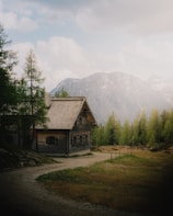 A rustic wooden cabin is nestled in a serene mountainous landscape, surrounded by lush green trees. The mountains in the background are partially covered with snow, and the sky above is filled with soft, fluffy clouds.