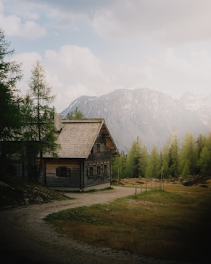 A rustic wooden cabin is nestled in a serene mountainous landscape, surrounded by lush green trees. The mountains in the background are partially covered with snow, and the sky above is filled with soft, fluffy clouds.