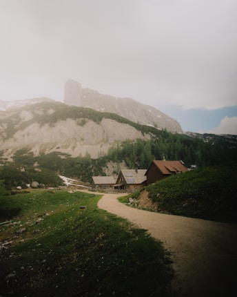 A serene mountain landscape features a misty hill with rugged cliffs rising above a dense forest. A winding dirt path leads to several rustic cabins with sloped roofs nestled at the base of the hill.