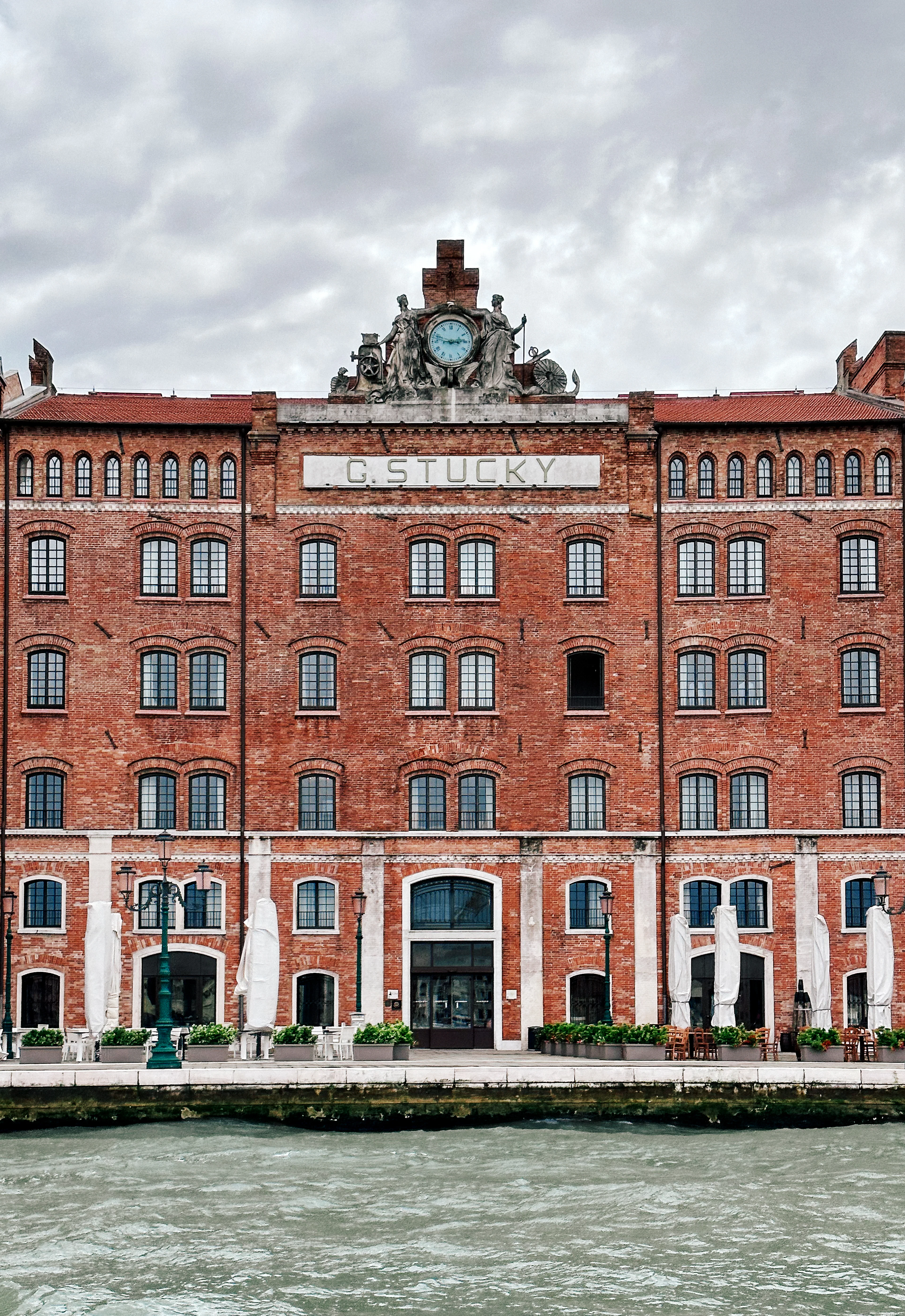 The red-brick neo-Gothic Molino Stucky on Giudecca in Venice, now a waterfront hotel, its clock and rooftop statuary crowning the facade above the lagoon and terrace tables