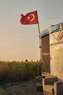 A Turkish flag waves atop a pole beside a rustic structure partially covered with a blue tarp. In the foreground, there is an empty armchair placed on the ground. The background features a field of sunflowers under a clear, warm sky.