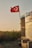 A Turkish flag waves atop a pole beside a rustic structure partially covered with a blue tarp. In the foreground, there is an empty armchair placed on the ground. The background features a field of sunflowers under a clear, warm sky.