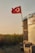 A Turkish flag waves atop a pole beside a rustic structure partially covered with a blue tarp. In the foreground, there is an empty armchair placed on the ground. The background features a field of sunflowers under a clear, warm sky.