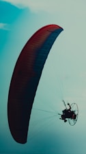 A paraglider silhouetted against a clear sky, with a large, brightly colored wing and a pilot seated in a harness. The scene conveys a sense of flight and adventure.