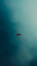 A single paraglider with a red canopy floats against a vast, cloudy sky. The scene captures a sense of freedom and solitude as the paraglider is framed by expansive blue hues.