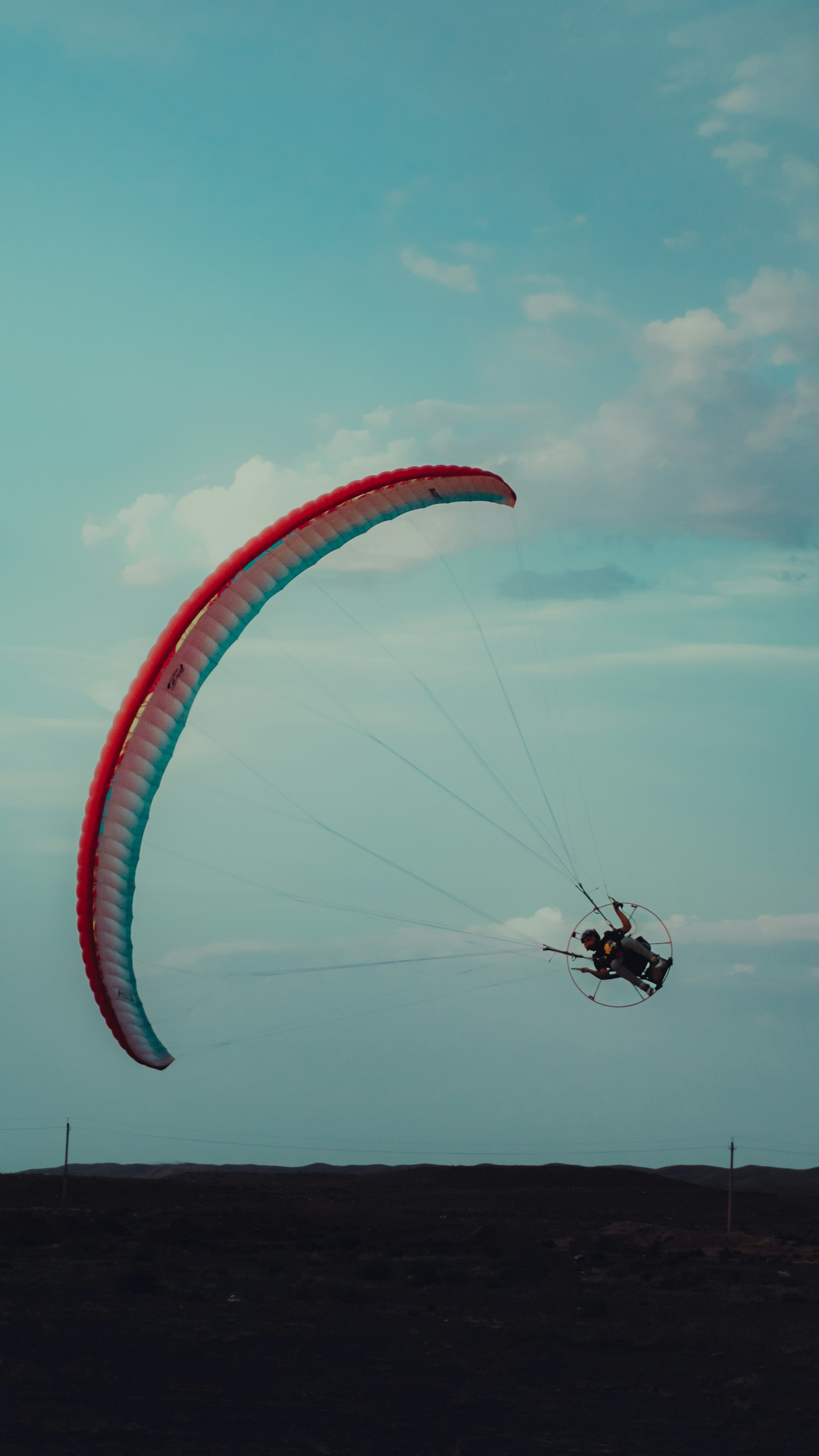 A paraglider with a large, colorful canopy soars through a clear sky, with subtle clouds visible. The paraglider is silhouetted against the evening sky, creating a serene and adventurous atmosphere.