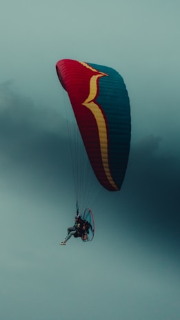 A paraglider with a colorful parachute canopy is soaring through a cloudy sky. The canopy has a vibrant pattern with red, blue, and yellow colors. The paraglider is wearing gear and is visible with legs slightly bent.