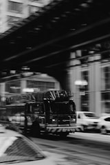 A fire truck is captured in motion on a city street with blurred surroundings, including buildings and other vehicles, indicating speed and urgency.