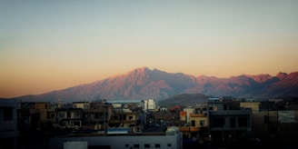 Residential buildings in the foreground with a scenic backdrop of rugged mountains bathed in the soft light of either sunrise or sunset.