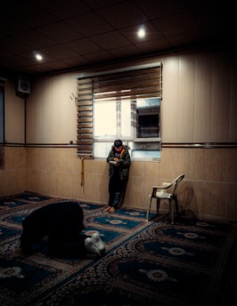 A dimly lit room with two people present. One person is kneeling on a patterned carpet in a position resembling prayer, while another stands by a window holding a book, leaning against the wall. The wooden walls and muted lighting create a serene and contemplative atmosphere. A plastic chair is positioned near the person by the window.