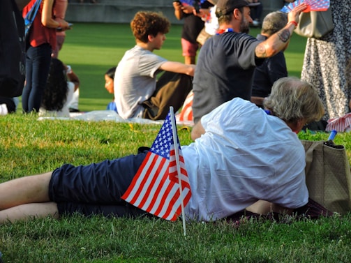 People are gathered outdoors on a grassy area, some sitting and some lying down, with a prominent American flag in the foreground. The group appears relaxed, suggesting a social or public event. Various individuals are holding small flags, adding to the patriotic theme.