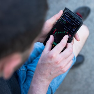 A person is using a smartphone to view a financial market chart. The screen displays a candlestick chart with green and red bars, likely indicating cryptocurrency or stock prices. The person appears to be seated, wearing casual clothes like shorts and holding the phone in both hands.