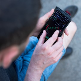 A person is using a smartphone to view a financial market chart. The screen displays a candlestick chart with green and red bars, likely indicating cryptocurrency or stock prices. The person appears to be seated, wearing casual clothes like shorts and holding the phone in both hands.