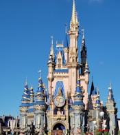 A majestic castle with blue and gold accents stands prominently against a clear blue sky. The structure is adorned with numerous towers and spires, and a large '50' emblem is prominently displayed on the front. Statues and colorful decorations are seen around the base.