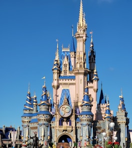 A majestic castle with blue and gold accents stands prominently against a clear blue sky. The structure is adorned with numerous towers and spires, and a large '50' emblem is prominently displayed on the front. Statues and colorful decorations are seen around the base.