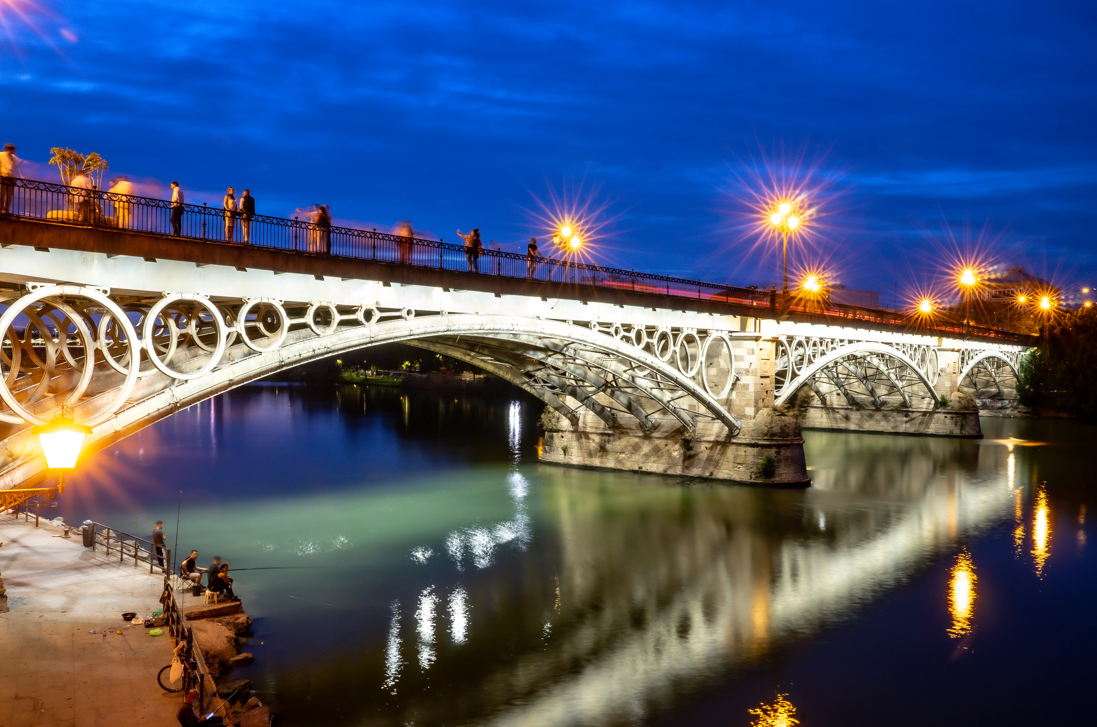 The Puente de Isabel II in Seville lit at blue hour, its white iron rings and lamps doubled in the Guadalquivir while people cross above and anglers sit on the quay.