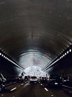 A congested traffic scene in a tunnel with vehicles lined up in lanes, heading towards the light at the tunnel's end. The tunnel walls and ceiling have a metallic, ribbed texture with rows of lights along the edges. The atmosphere is dimly lit, creating a contrast with the bright exit ahead.
