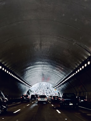 A congested traffic scene in a tunnel with vehicles lined up in lanes, heading towards the light at the tunnel's end. The tunnel walls and ceiling have a metallic, ribbed texture with rows of lights along the edges. The atmosphere is dimly lit, creating a contrast with the bright exit ahead.