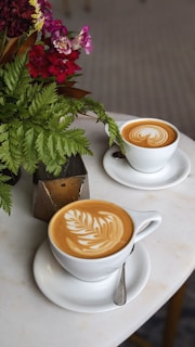 Two cups of latte art coffee are placed on a white table, accompanied by a decorative plant arrangement with green leaves and red and purple flowers. The coffee cups are white, with intricate designs created in the foam. A brown geometric vase or container is also present next to the plant.