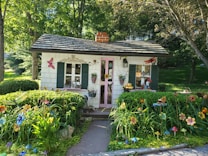 A quaint, small cottage with a shingled roof and pink door is nestled in a lush garden. The cottage is adorned with shutters and decorative elements like butterfly and flowers. The surrounding area is vibrant with greenery and colorful flowering plants, creating a whimsical and inviting atmosphere.