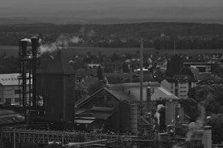 An industrial facility with several large structures and smokestacks emitting plumes of smoke. The background features a landscape of trees and a small residential area. Overcast skies add to the somber mood.