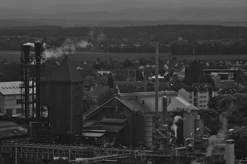An industrial facility with several large structures and smokestacks emitting plumes of smoke. The background features a landscape of trees and a small residential area. Overcast skies add to the somber mood.