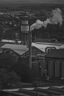 A black and white photograph depicting an industrial landscape with a prominent smokestack emitting white smoke into the sky. Surrounding the smokestack are several large industrial buildings and storage tanks. In the background, a town and a forested area are visible.