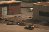 A construction site features a large dirt area with a yellow loader parked in the foreground. The background includes two industrial buildings, one made of brick and the other with metal siding. A white van is parked near a fence that separates the site from the road. Several barriers and a pile of gravel and dirt can be seen around the site.