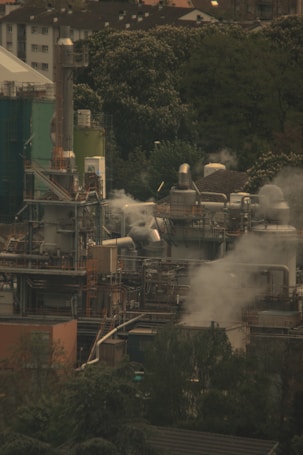 An industrial complex with numerous metal structures, chimneys, and storage tanks emitting steam surrounded by dense greenery and residential buildings in the background.