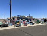 A vibrant and eclectic outdoor installation made up of numerous painted television sets stacked and arranged on a gravel section beside a street. The television sets feature various designs, patterns, and faces painted in bright colors, adding an artistic touch to the area. Surrounding buildings include a small house with an American flag visible and additional structures under the clear blue sky.