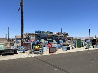 A vibrant and eclectic outdoor installation made up of numerous painted television sets stacked and arranged on a gravel section beside a street. The television sets feature various designs, patterns, and faces painted in bright colors, adding an artistic touch to the area. Surrounding buildings include a small house with an American flag visible and additional structures under the clear blue sky.