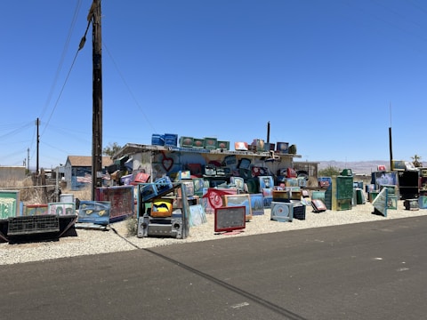 A vibrant and eclectic outdoor installation made up of numerous painted television sets stacked and arranged on a gravel section beside a street. The television sets feature various designs, patterns, and faces painted in bright colors, adding an artistic touch to the area. Surrounding buildings include a small house with an American flag visible and additional structures under the clear blue sky.