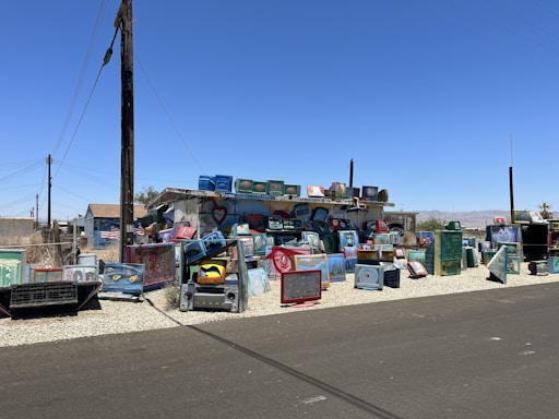 A vibrant and eclectic outdoor installation made up of numerous painted television sets stacked and arranged on a gravel section beside a street. The television sets feature various designs, patterns, and faces painted in bright colors, adding an artistic touch to the area. Surrounding buildings include a small house with an American flag visible and additional structures under the clear blue sky.