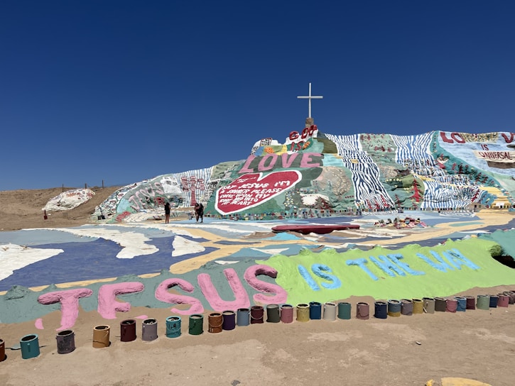 A colorful hillside covered with vibrant, hand-painted religious and inspirational messages. A cross is prominently placed at the top of the hill. The foreground features large painted letters spelling out 'JESUS IS THE WAY,' surrounded by paint cans. The central part of the hill displays the word 'LOVE' in large letters, with a heart shape underneath.