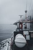 A boat navigating through calm waters on a foggy day is captured. The deck features a cylindrical, white object secured with ropes, likely a life raft or buoy. A small radar or antenna is mounted above a cabin surrounded by various equipment and safety signage. The horizon is obscured by mist, adding a sense of mystery.