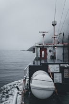 A boat navigating through calm waters on a foggy day is captured. The deck features a cylindrical, white object secured with ropes, likely a life raft or buoy. A small radar or antenna is mounted above a cabin surrounded by various equipment and safety signage. The horizon is obscured by mist, adding a sense of mystery.