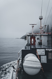 A boat navigating through calm waters on a foggy day is captured. The deck features a cylindrical, white object secured with ropes, likely a life raft or buoy. A small radar or antenna is mounted above a cabin surrounded by various equipment and safety signage. The horizon is obscured by mist, adding a sense of mystery.