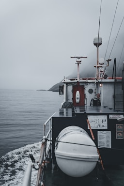 A boat navigating through calm waters on a foggy day is captured. The deck features a cylindrical, white object secured with ropes, likely a life raft or buoy. A small radar or antenna is mounted above a cabin surrounded by various equipment and safety signage. The horizon is obscured by mist, adding a sense of mystery.