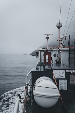 A boat navigating through calm waters on a foggy day is captured. The deck features a cylindrical, white object secured with ropes, likely a life raft or buoy. A small radar or antenna is mounted above a cabin surrounded by various equipment and safety signage. The horizon is obscured by mist, adding a sense of mystery.