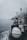 A boat navigating through calm waters on a foggy day is captured. The deck features a cylindrical, white object secured with ropes, likely a life raft or buoy. A small radar or antenna is mounted above a cabin surrounded by various equipment and safety signage. The horizon is obscured by mist, adding a sense of mystery.