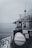 A boat navigating through calm waters on a foggy day is captured. The deck features a cylindrical, white object secured with ropes, likely a life raft or buoy. A small radar or antenna is mounted above a cabin surrounded by various equipment and safety signage. The horizon is obscured by mist, adding a sense of mystery.