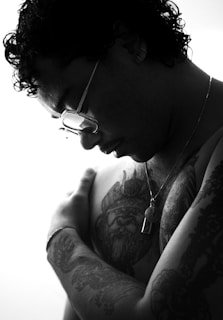 A person with curly hair is depicted in a black and white image, looking downward and embracing themselves. They are wearing glasses, earrings, and a necklace with a pendant. Tattoos are prominently visible on their arm and chest, including a detailed design on the chest.