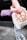 A person is holding a light-colored bag with peace symbols and the word 'Love.' The bag also contains a purple yoga mat. The individual is dressed in a pink long-sleeve shirt and floral-patterned leggings. The ground is made of gray pavement tiles, and there is a dark wall in the background.