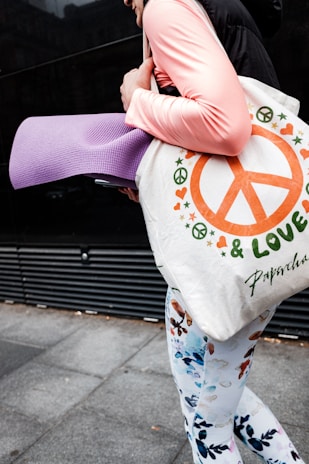 A person is holding a light-colored bag with peace symbols and the word 'Love.' The bag also contains a purple yoga mat. The individual is dressed in a pink long-sleeve shirt and floral-patterned leggings. The ground is made of gray pavement tiles, and there is a dark wall in the background.