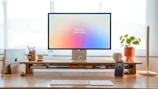 A minimalistic desk setup featuring a large monitor with the text 'think less, create more.' There are various items organized neatly including a laptop, a smartphone on a stand, a potted plant, and decorative figures. The desk is made of light wood, and natural light is coming through the window in the background.