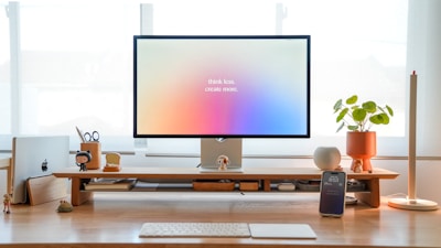 A minimalistic desk setup featuring a large monitor with the text 'think less, create more.' There are various items organized neatly including a laptop, a smartphone on a stand, a potted plant, and decorative figures. The desk is made of light wood, and natural light is coming through the window in the background.