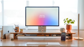 A minimalistic desk setup featuring a large monitor with the text 'think less, create more.' There are various items organized neatly including a laptop, a smartphone on a stand, a potted plant, and decorative figures. The desk is made of light wood, and natural light is coming through the window in the background.
