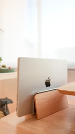 A silver laptop is placed vertically on a wooden laptop stand. The stand is made out of light-colored wood and has a sleek, minimalist design. The setting includes a soft, bright background that suggests natural lighting, and possibly some indoor plants or decorations nearby.